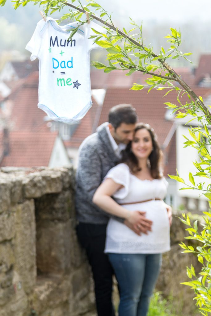 Schwangeres Paar an einer Mauer, an einem Zweig hängt ein Babybody