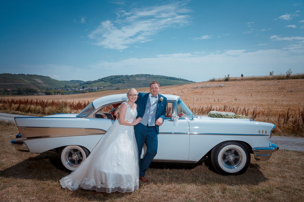 Brautpaar an einem hellen Oldtimer vor Feldern und Hügeln