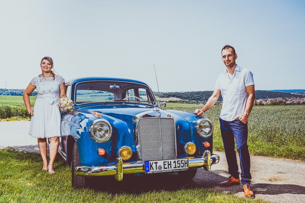 Frau und Mann vor einem blauen Oldtimer auf einer Wiese