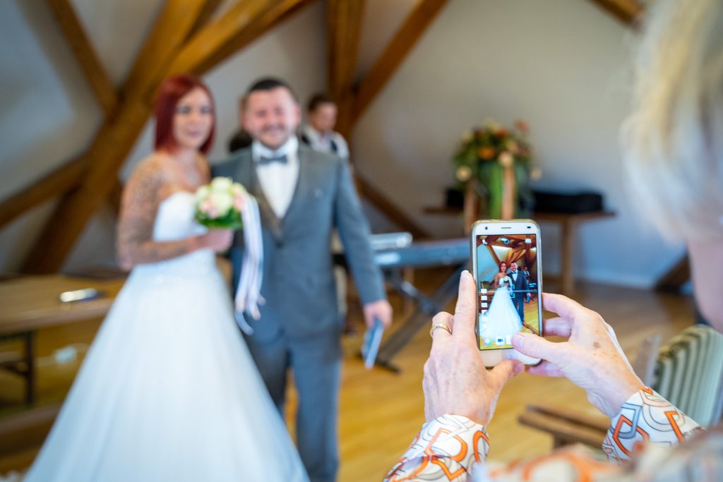 Brautpaar in einem Saal mit Holzbalken, im Vordergrund fotografiert jemand mit dem Handy